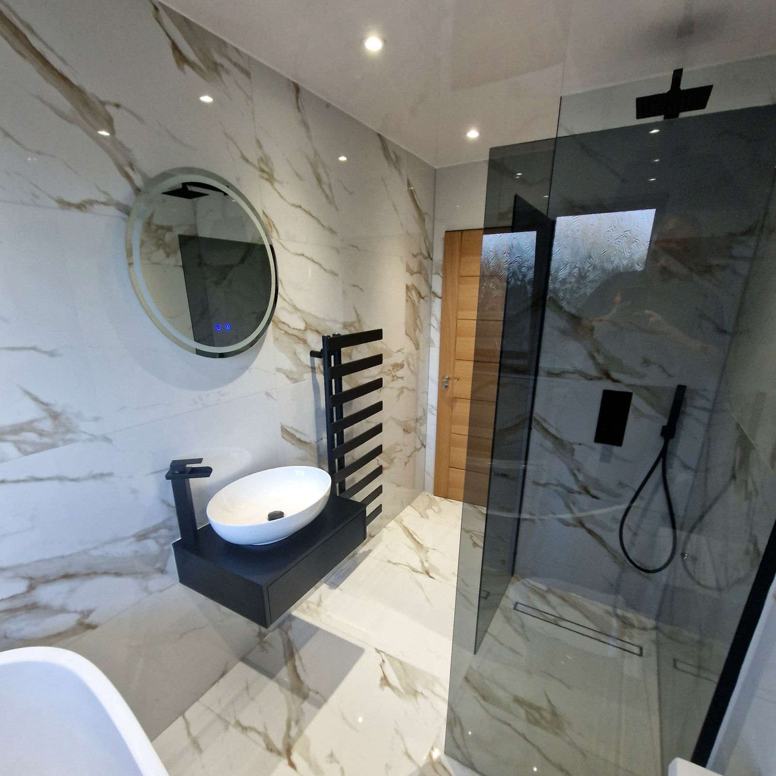 A backlit round mirror over a white vessel basin on a dark floating shelf, beside a smoked glass shower screen