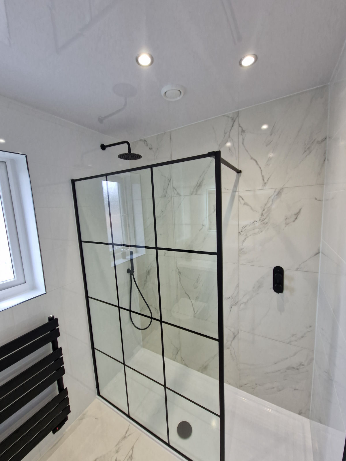 A black grid shower screen over a bath, against full-height marble-effect tiling
